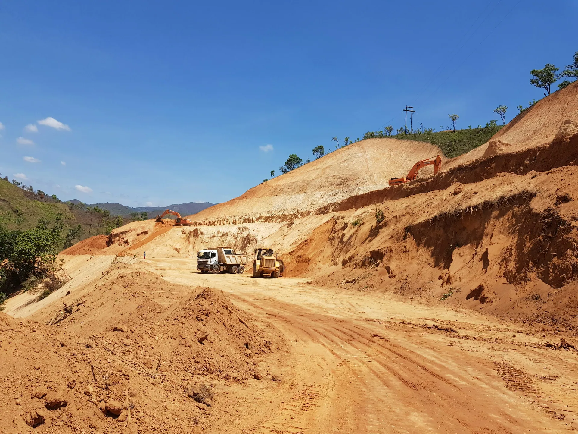 Dump truck, loader and excavator working a red-soil hillside cut under blue sky